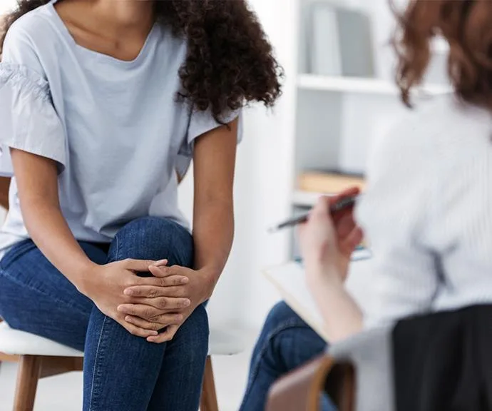 Person sitting cross-legged, holding hands in a therapy session, with another person writing notes in the background.