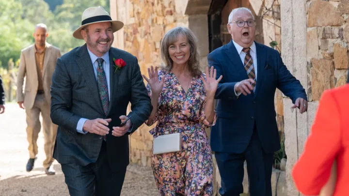 A group of people in formal wear smiling and walking outside, one man wears a hat with a red rose boutonnière.