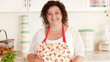 Smiling woman in a kitchen wearing a strawberry-patterned apron, with jars and greenery on the counter.