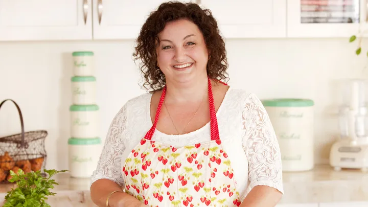 Smiling woman in a kitchen wearing a strawberry-patterned apron, with jars and greenery on the counter.