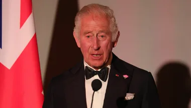 Man in a tuxedo speaking at a podium, with a Union Jack flag in the background.