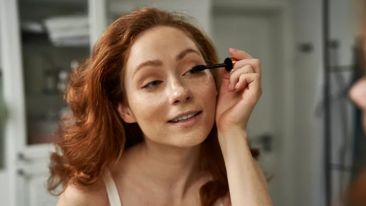 A woman with red hair applying mascara in front of a mirror.