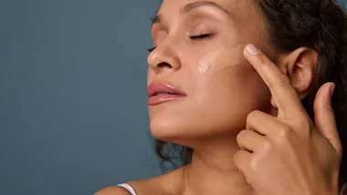 A woman applying foundation to her cheek with her finger, eyes closed, against a blue background.
