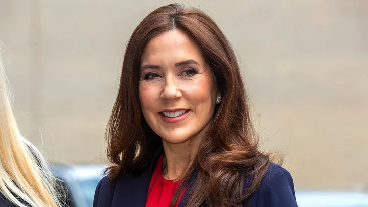 A woman with long brown hair is smiling and wearing a red top and navy jacket.