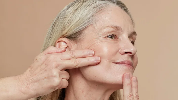 Older woman gently pressing her cheek with fingers, showcasing skin texture and lines.