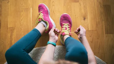 Person tying yellow laces on pink running shoes, wearing teal leggings on a wooden floor.