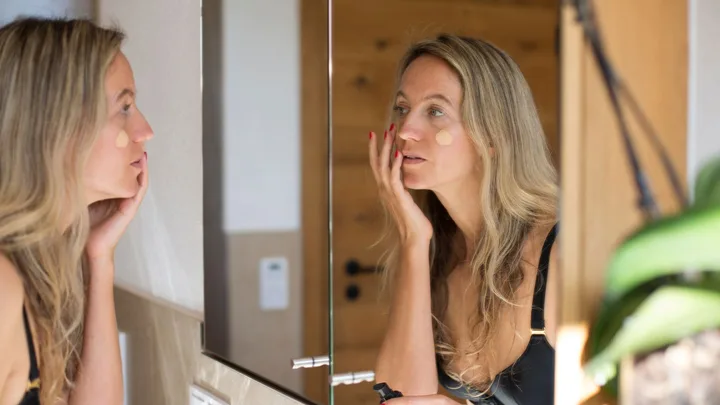 Woman applying foundation in front of a bathroom mirror, with focus on her reflection.