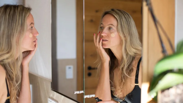 Woman applying foundation in front of a bathroom mirror, with focus on her reflection.