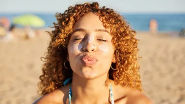 Woman with curly hair and sunscreen poses playfully, puckering lips at a sunny beach.