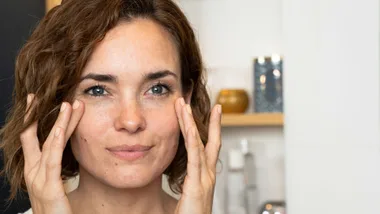 Woman applying skincare under eyes, smiling softly, blurred background with shelves.