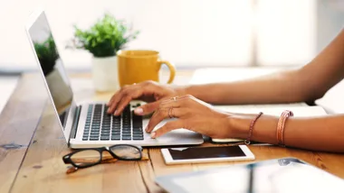 Person typing on a laptop at a wooden table with a smartphone, glasses, notebook, and yellow mug nearby.