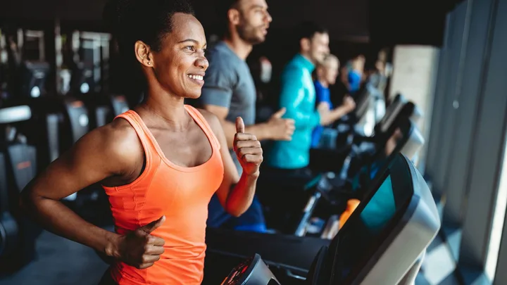 People exercising on treadmills at a gym, with a woman in an orange tank top in the foreground.
