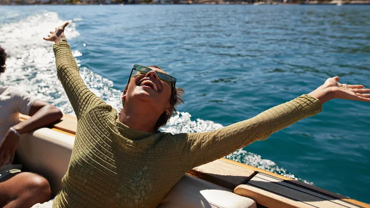 Person joyfully stretching arms wide on a boat, wearing sunglasses, with clear blue water in the background.