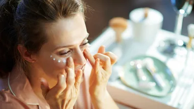Woman applying skincare cream under eyes at a vanity table.