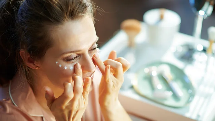 Woman applying skincare cream under eyes at a vanity table.