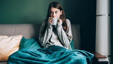 Woman in a cozy sweater and blanket drinking from a mug on a couch with teal pillows.
