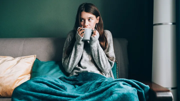 Woman in a cozy sweater and blanket drinking from a mug on a couch with teal pillows.