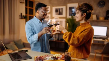 Two people clink glasses of red wine in a cozy, dimly lit living room with a cheese platter and laptop on the table.