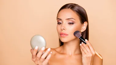 Woman applying makeup with a brush while holding a compact mirror against a peach background.