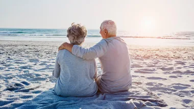 Elderly couple sitting on a beach, wrapped in a blanket, watching the sunset over the ocean.