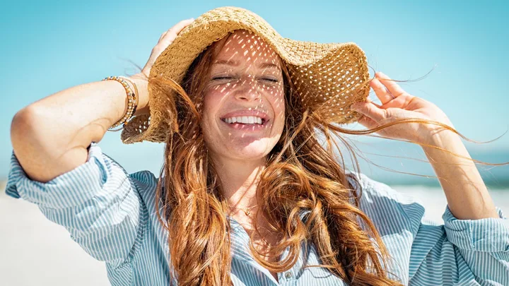 Smiling woman on a sunny beach wearing a straw hat and striped shirt, with windblown red hair.