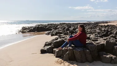 A woman sits on rocks by a sunny beach, gazing at the ocean.