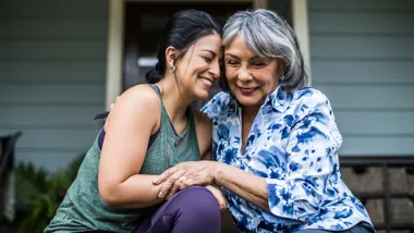 Smiling older woman and younger woman embrace warmly on a porch step.