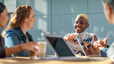 Group of diverse people in a meeting discussing and smiling, with laptops and coffee cups on the table.