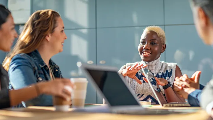 Group of diverse people in a meeting discussing and smiling, with laptops and coffee cups on the table.
