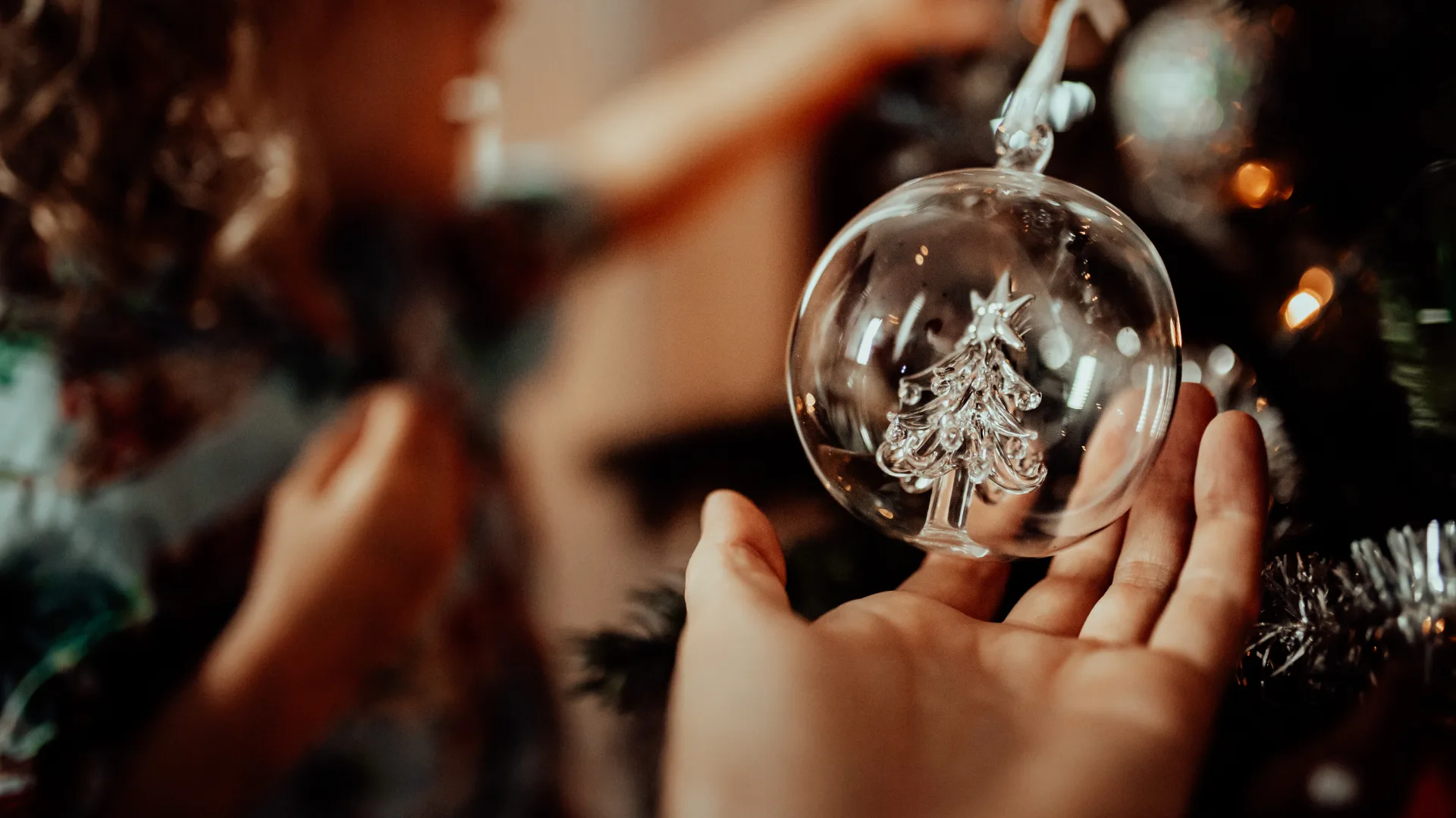 Hand holding a clear ornament with a small tree inside, while a child decorates a Christmas tree in the background.