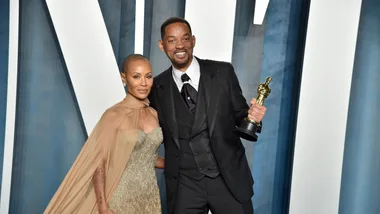 A man in a black suit holds an Oscar statue, posing with a woman in a gold dress on a blue and white backdrop.