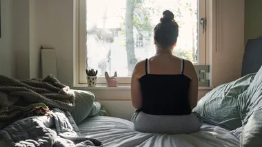 Person sitting on bed facing a window with soft morning light coming in; cozy blankets and potted plants nearby.