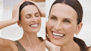 Smiling woman with hands on head and cheek, wearing hoop earrings, long brown hair pulled back.