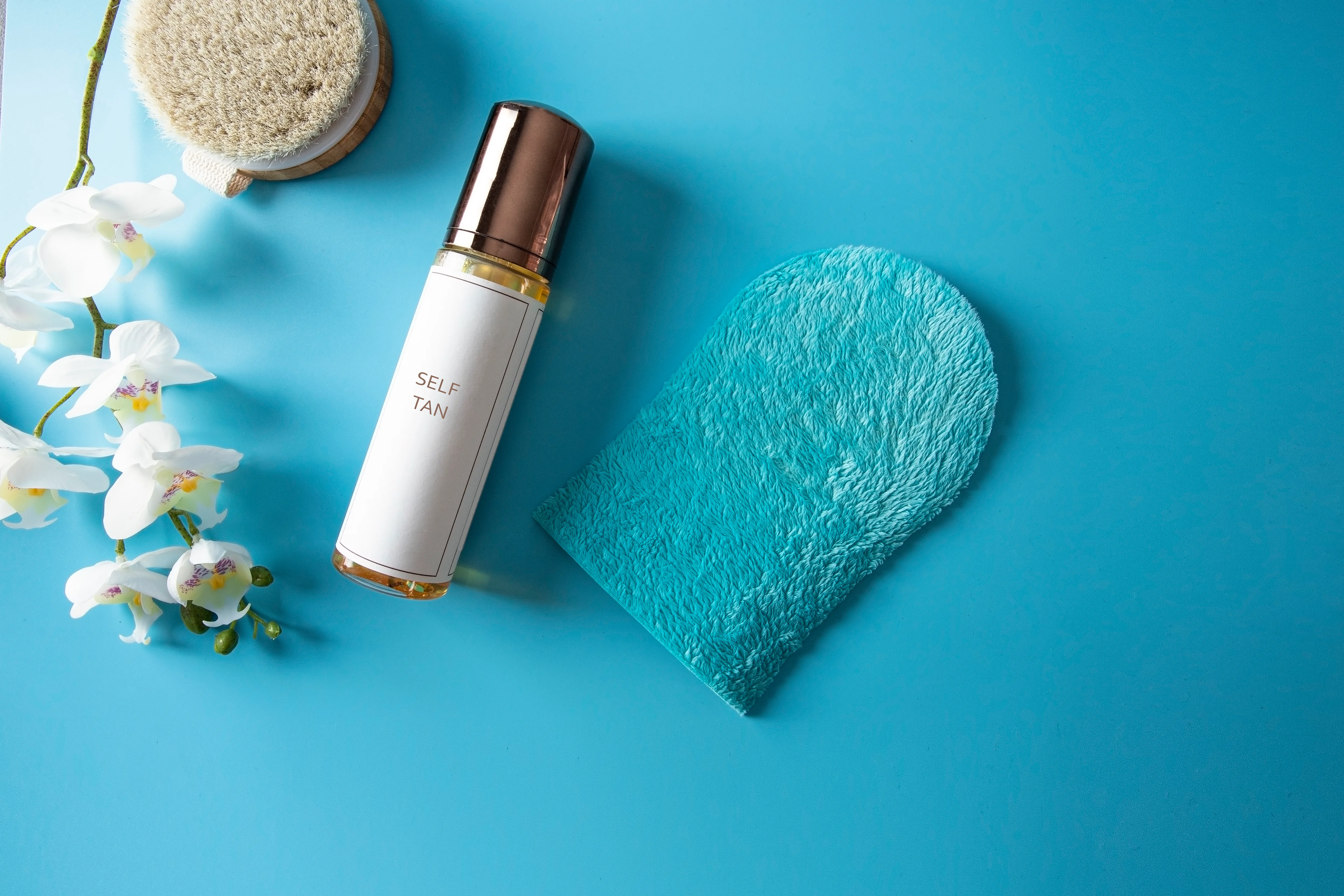 Gradual self tanning concept. Exfoliating Skin Brush, self tan lotion and  tanning mitt on blue background with orchid flower .