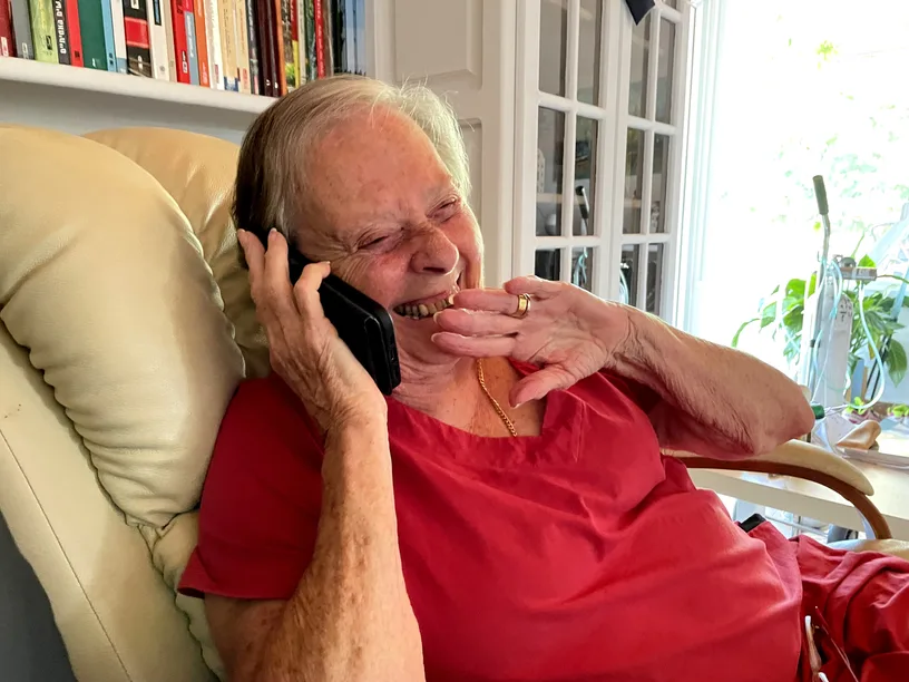 Roni, at home in Israel, is on the phone with Rami. She is wearing a red top and she is laughing.