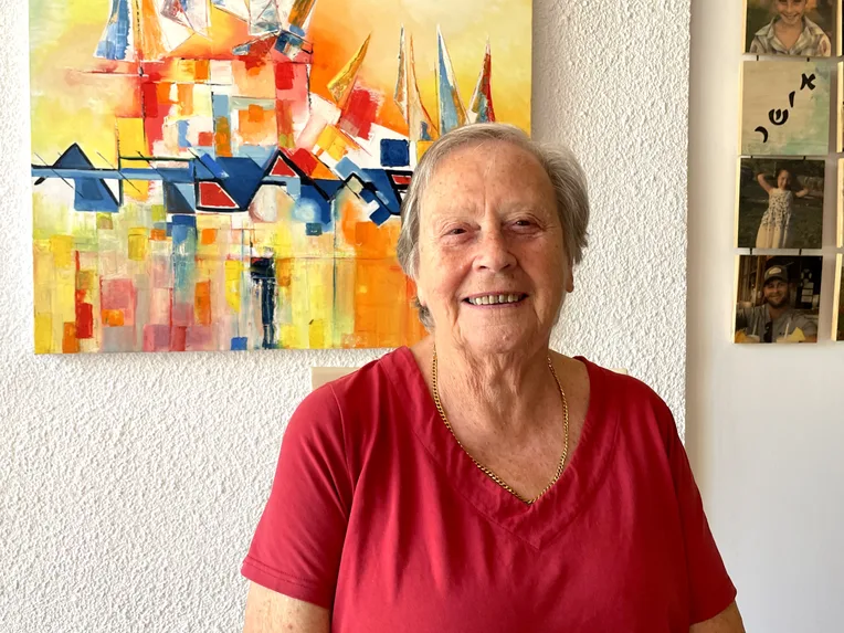 Roni is wearing a red top and smiling. She is at home in Israel, standing in front of a colourful painting.