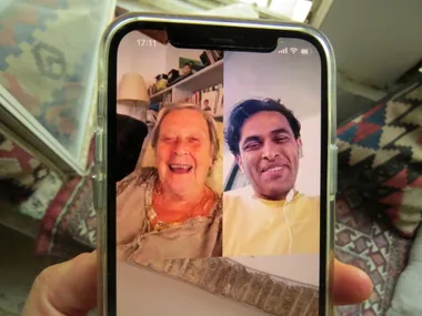 Video call on a smartphone showing two people smiling, held by a person in a cozy room setting.