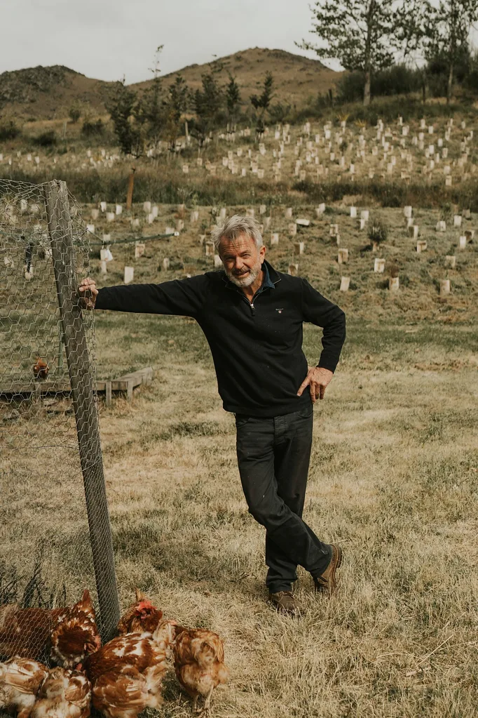 Man leaning on a chicken coop fence in a hilly field with chickens nearby.