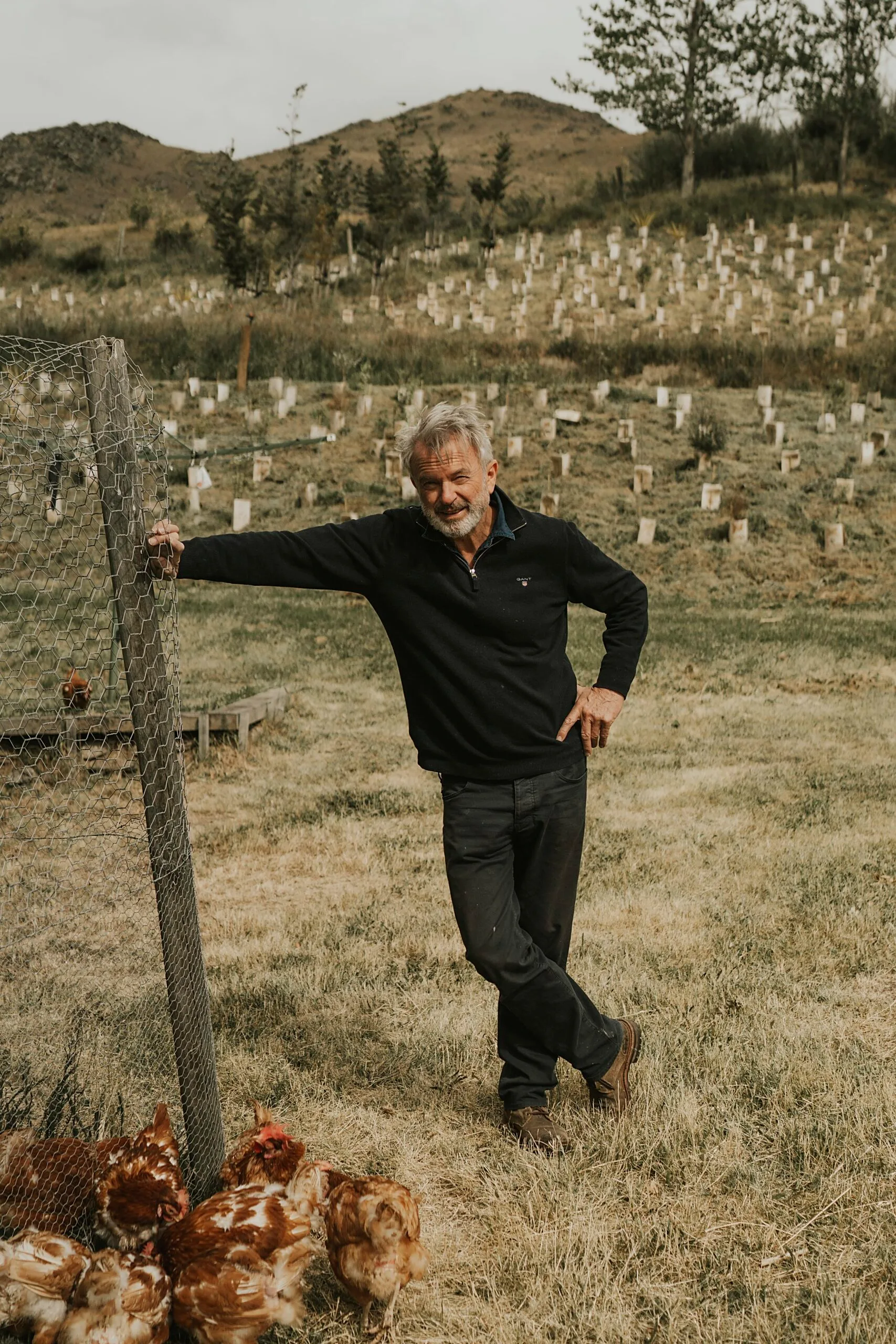 Man leaning on a chicken coop fence in a hilly field with chickens nearby.