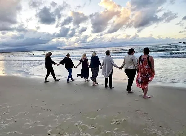 Seven women standing on a beach, looking out to sea. 