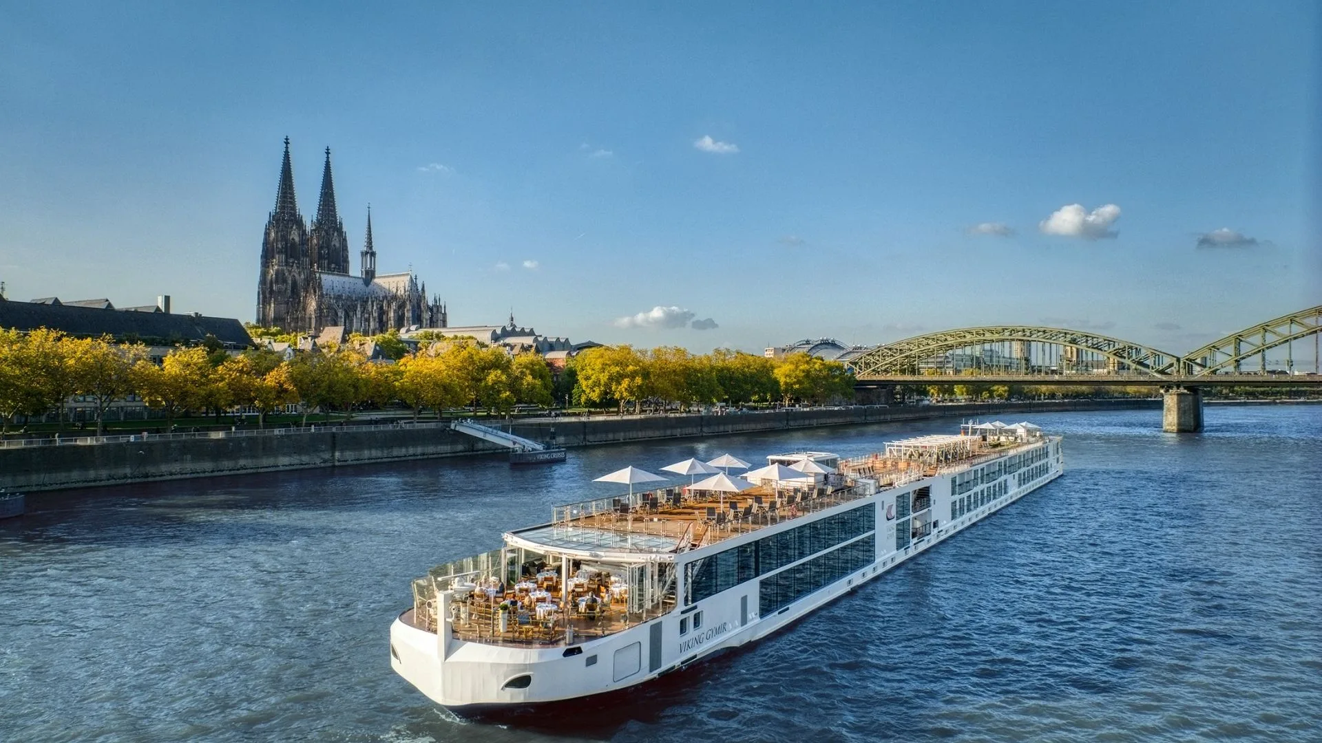 River cruise ship near Cologne Cathedral and Hohenzollern Bridge on the Rhine with autumn trees and clear sky.