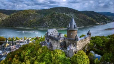 Aerial view of Stahleck Castle by the Rhine River with surrounding hills and a cruise ship sailing.