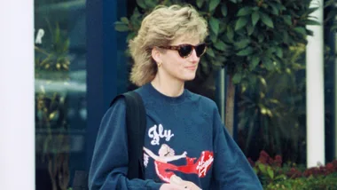 A woman wearing sunglasses and a casual sweatshirt with "Fly" design, walking outside with plants in the background.