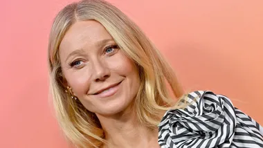 A person with long blonde hair smiles against a pink background, wearing a striped black and white top.