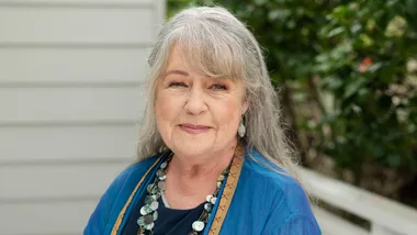 Elderly woman with long gray hair smiling, wearing a blue top and statement necklace, outdoors with greenery in background.