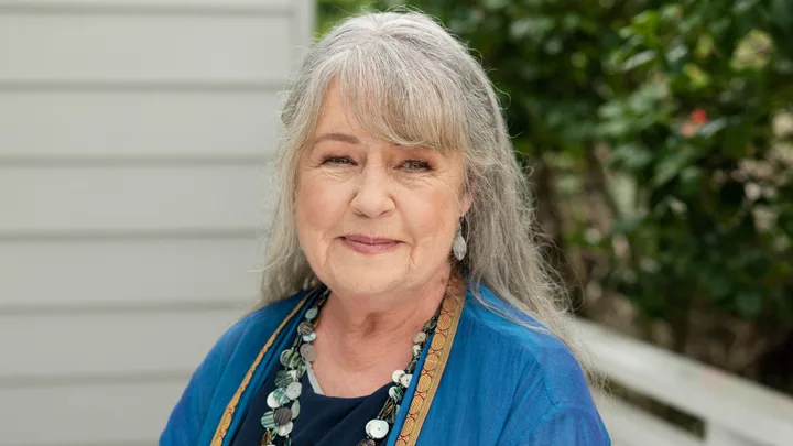 Elderly woman with long gray hair smiling, wearing a blue top and statement necklace, outdoors with greenery in background.