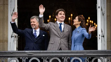 Three people waving from a balcony, wearing formal attire with a chandelier in the background.