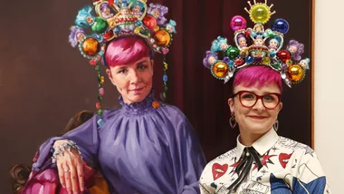 A woman with pink hair and colorful, doll-adorned headpiece stands beside a painting of herself wearing the same attire.