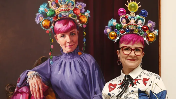 A woman with pink hair and colorful, doll-adorned headpiece stands beside a painting of herself wearing the same attire.