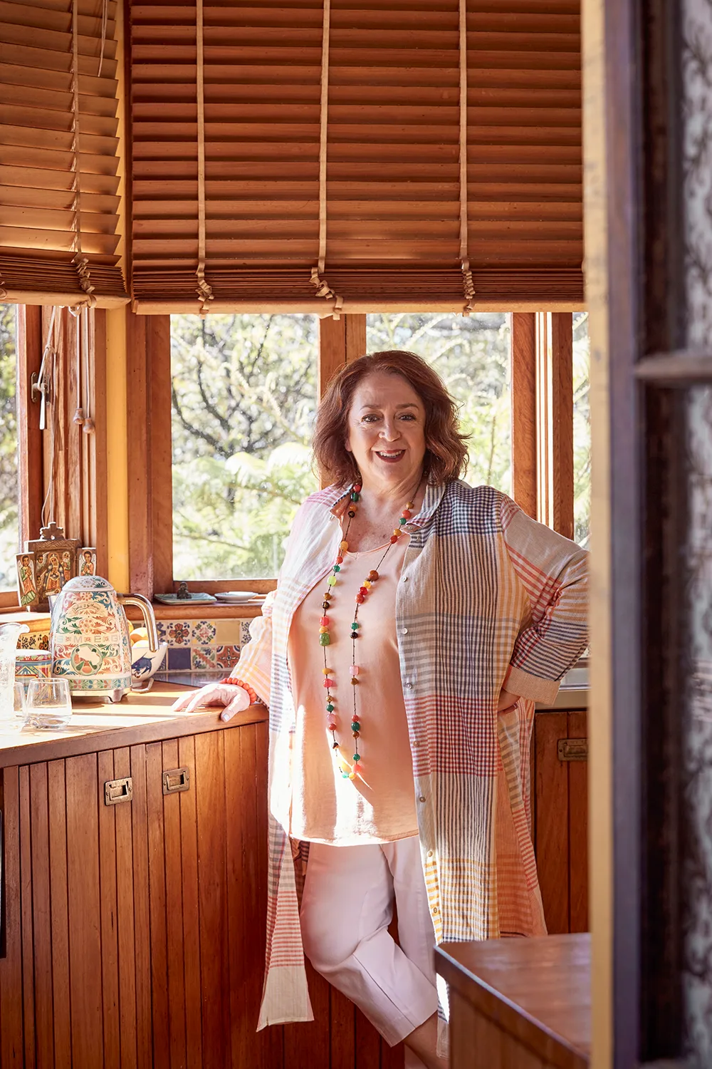 Wendy Harmer is standing at her kitchen bench with a window behind her. She is wearing a long, colourful, checked shirt and beads.